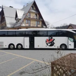 Biały autokar z otwartymi drzwiami, zaparkowany na brukowanym parkingu przed budynkiem w stylu góralskim z szarym dachem. Na boku autobusu widoczne logo firmy.