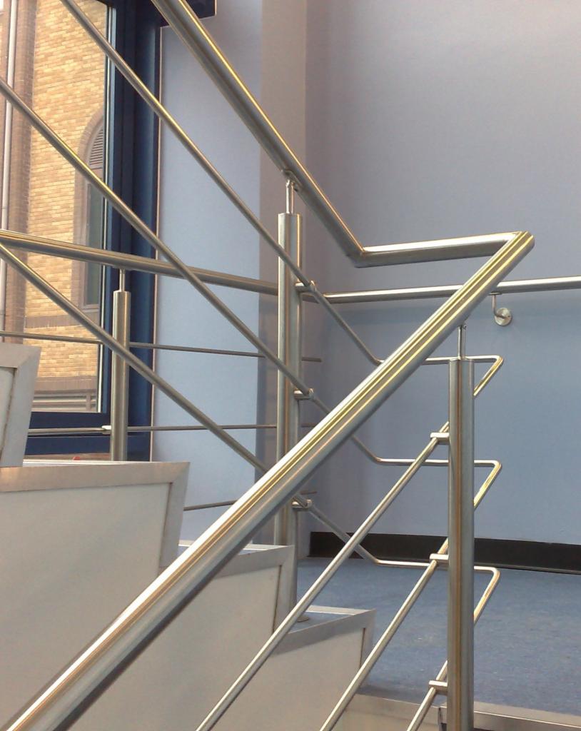 Nowoczesne, metalowe barierki schodowe z elementami ze stali nierdzewnej. Widok z dołu, klatka schodowa, okno w tle.