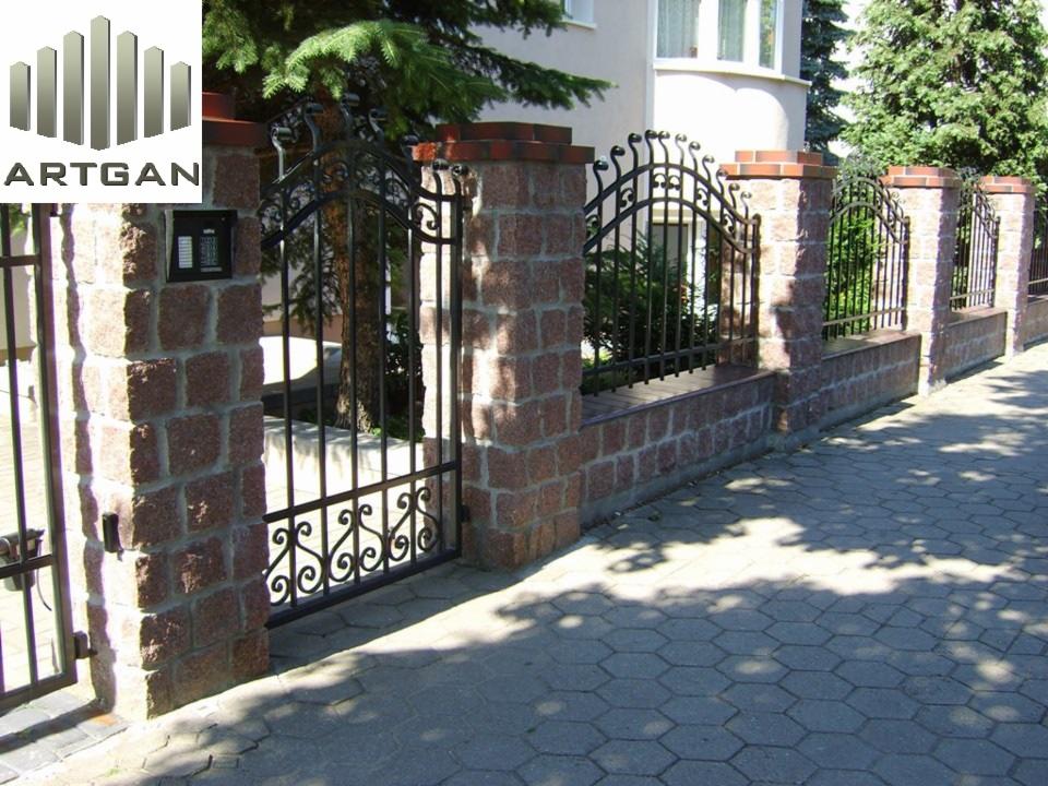 Kute ogrodzenie z bramą, murowane słupki z czerwonej cegły, dekoracyjne elementy metalowe. Widok na posesję i chodnik z kostki brukowej.