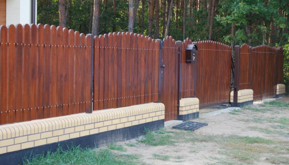 Brunatne ogrodzenie drewniane z furtką i skrzynką na listy, osadzone na niskiej, ceglanej podmurówce, na tle leśnego krajobrazu. Widoczne nity łączące deski.