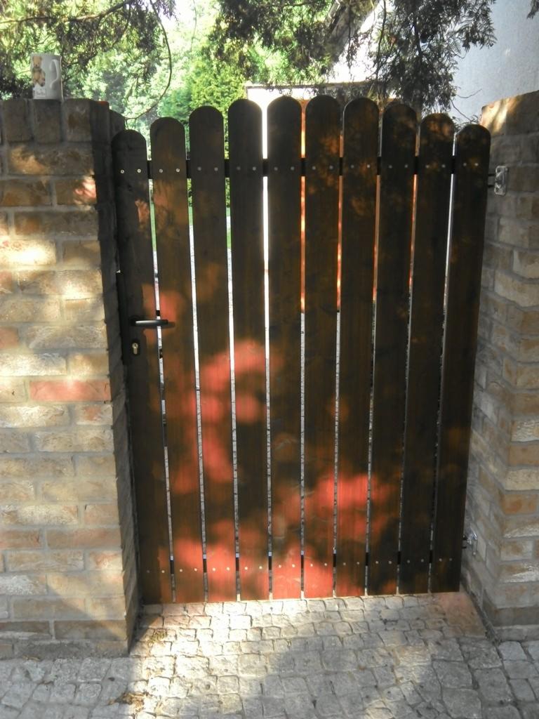 Ciemnobrązowa furtka z pionowych desek, osadzona w ceglanych słupkach, z widocznymi promieniami słońca i kostką brukową u dołu. Furtka zamyka wejście na posesję.