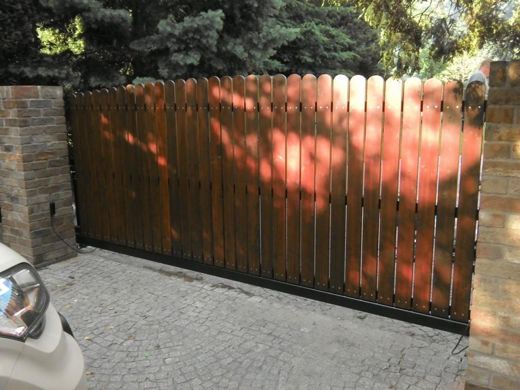 Drewniana brama przesuwna z pionowymi deskami, osadzona między murowanymi słupkami. Widoczne cienie drzew i fragment samochodu po lewej stronie.