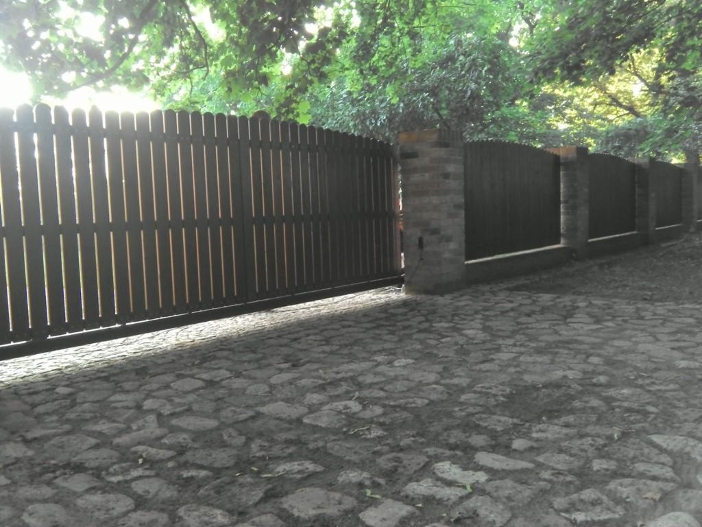 Ciemne, drewniane ogrodzenie panelowe z bramą wjazdową, osadzone na niskiej podmurówce i ceglanych słupkach, na tle zieleni drzew i kamiennej nawierzchni.