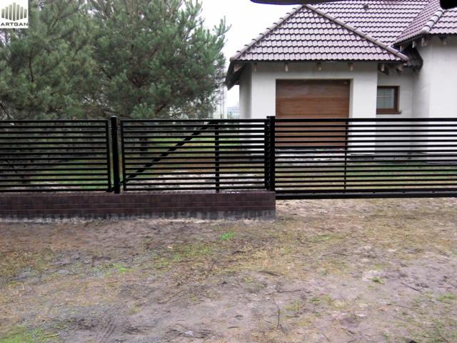 Nowoczesne, poziome ogrodzenie metalowe w kolorze czarnym, osadzone na ceglanej podmurówce. W tle dom jednorodzinny z garażem i zielone drzewa.
