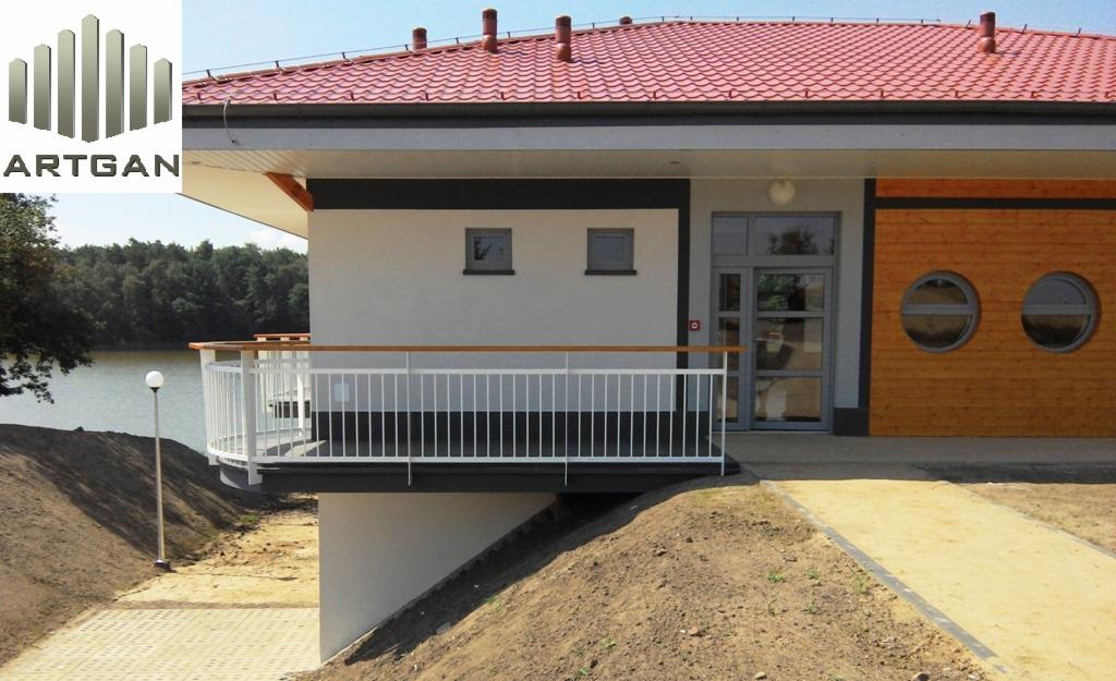 Nowoczesne ogrodzenie balkonowe z białymi, pionowymi prętami i drewnianą poręczą, z widokiem na jezioro, przylegające do budynku z drewnianą elewacją.