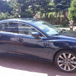 Błękitna Mazda 6 sedan zaparkowana na brukowanym podjeździe, widok z boku na tle zieleni.