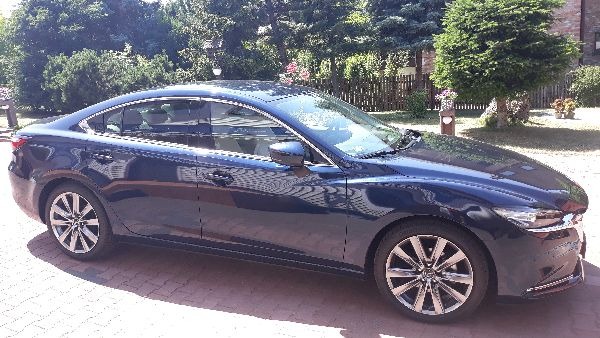 Błękitna Mazda 6 sedan zaparkowana na brukowanym podjeździe, widok z boku na tle zieleni.