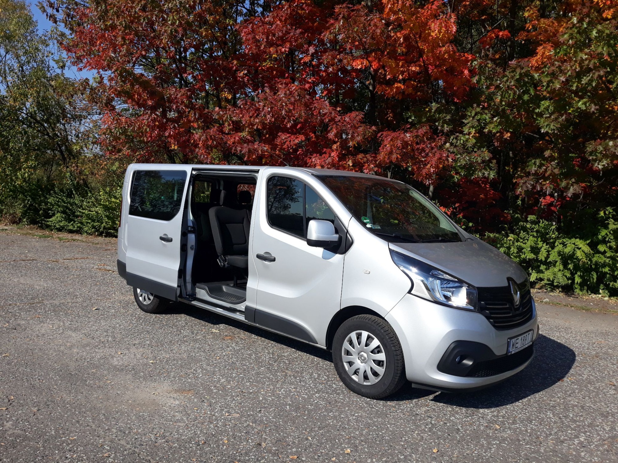 Srebrny van Renault Trafic z otwartymi drzwiami przesuwnymi, zaparkowany na szutrowej drodze, w tle drzewa z jesiennymi liśćmi.