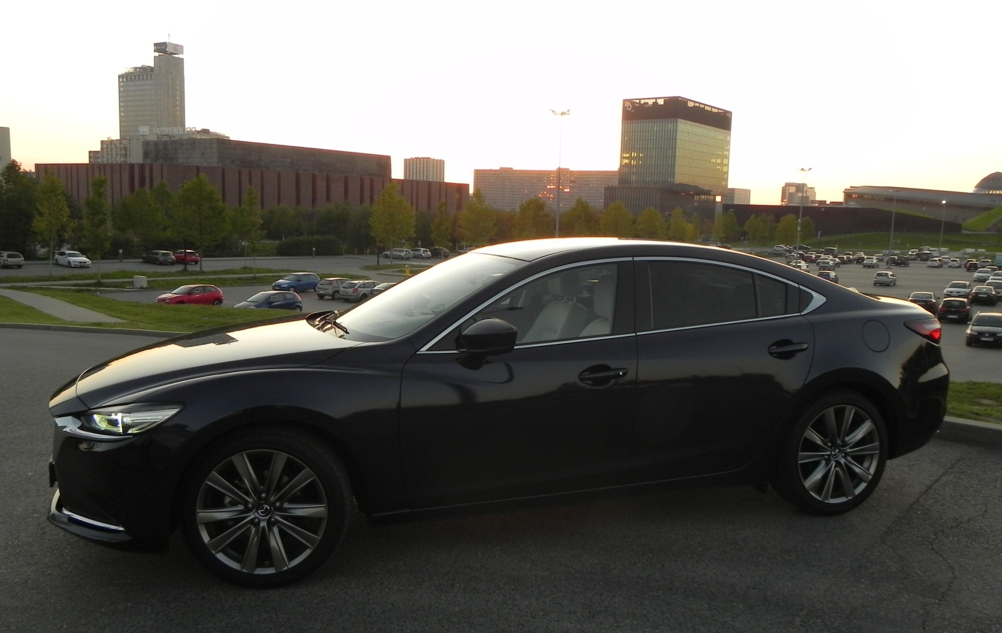 Czarny samochód osobowy Mazda 6 na parkingu miejskim z budynkami w tle, widok z boku.