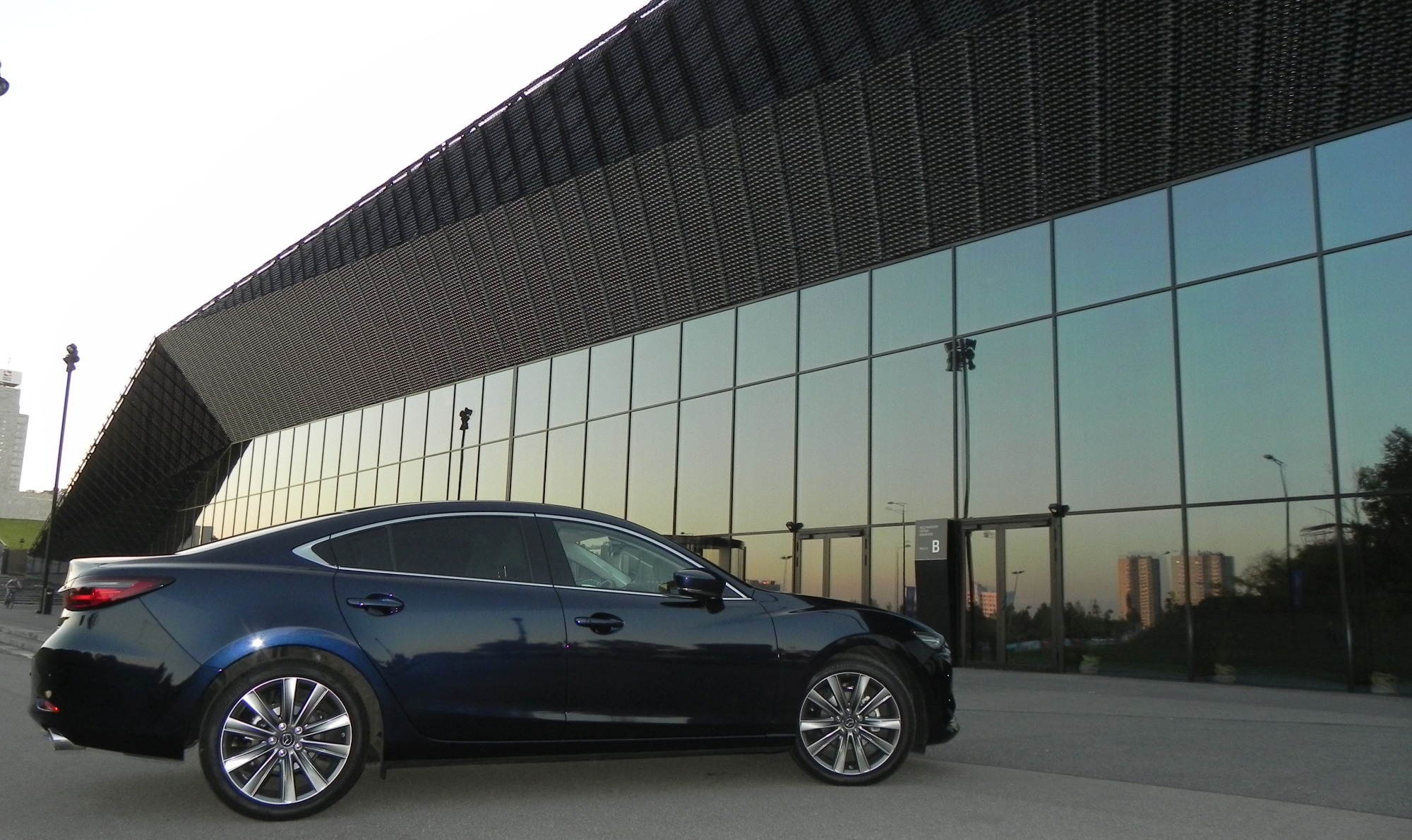 Granatowy sedan Mazda zaparkowany przed nowoczesnym budynkiem z lustrzanymi oknami, odbijającymi zachodzące słońce.
