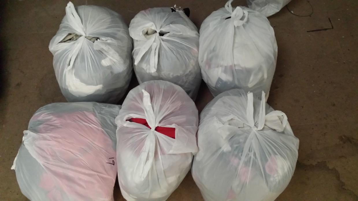 Sześć worków foliowych wypełnionych odzieżą, widoczne różne kolory materiałów, leżące na podłodze. Jeden z worków ma numer '19' napisany markerem.