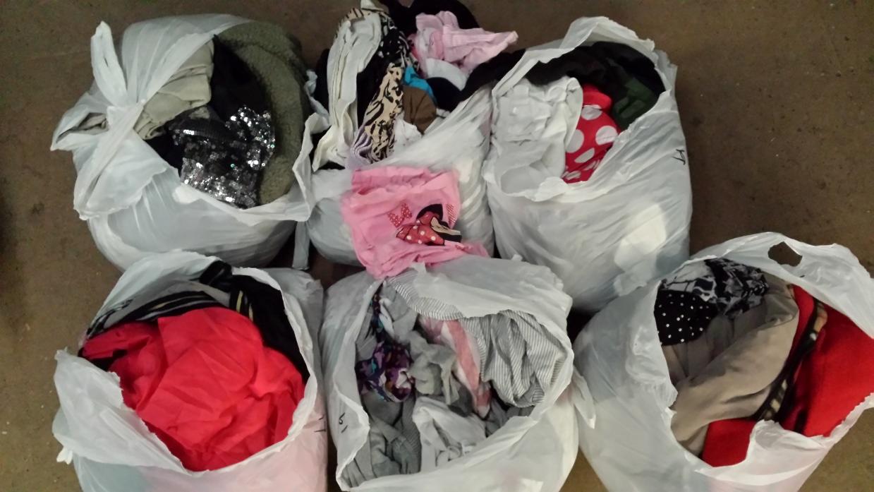 Sześć worków foliowych wypełnionych różnorodną odzieżą używaną, w tym bluzki w kropki, cekiny i ubrania w paski. Część ubrań wystaje z worków.