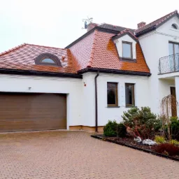 Dwukondygnacyjny dom z czerwonym dachem i brązową bramą garażową, otoczony zielenią. Widoczny balkon z ozdobną balustradą i podjazd z kostki brukowej.