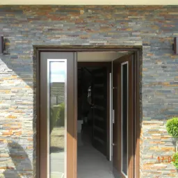 Wejście do domu z elewacją z kamienia dekoracyjnego w odcieniach szarości i brązu, oświetlone dwoma kinkietami. Drzwi wejściowe drewniane z przeszkleniami, obok drzwi dwie małe, formowane rośliny.