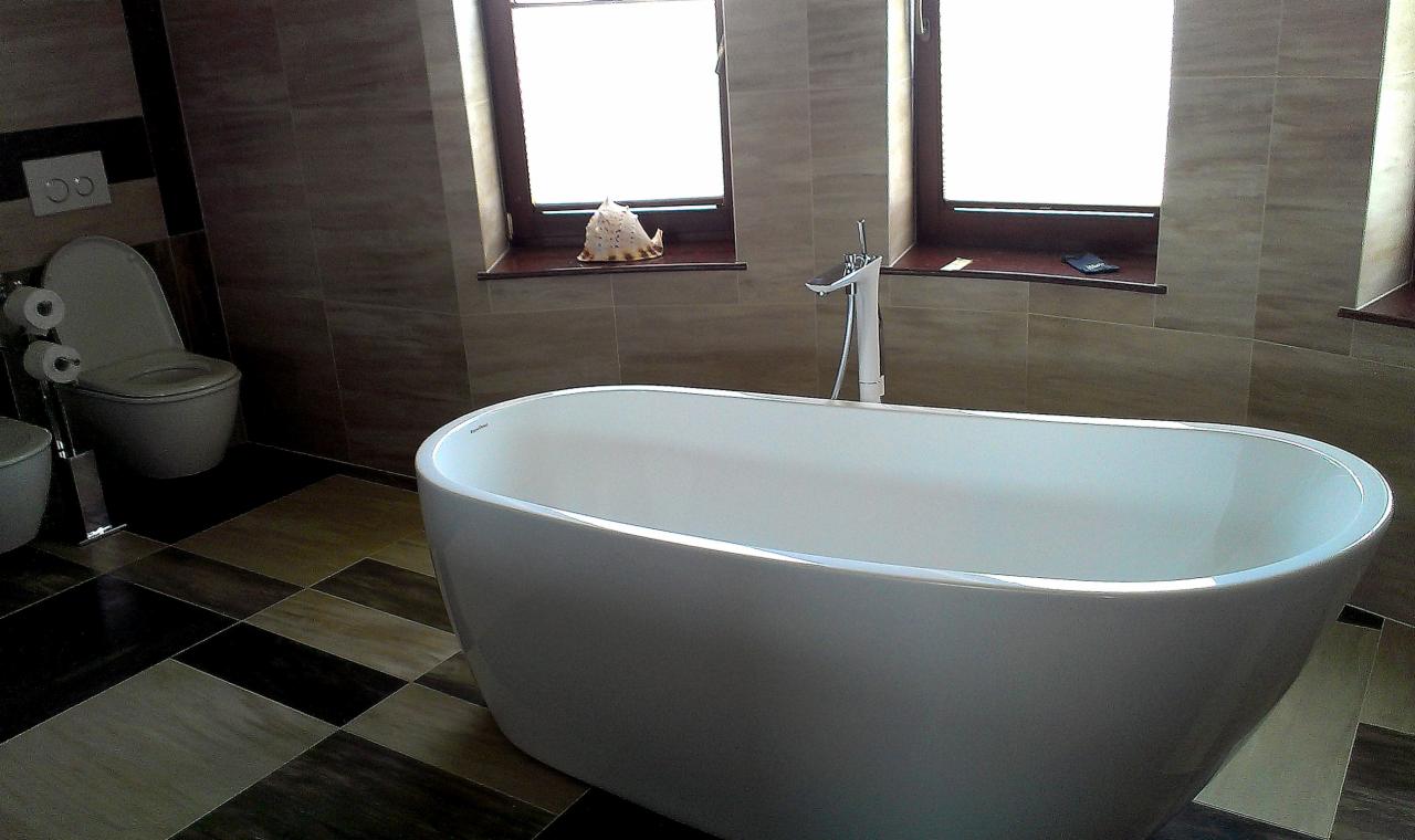 Nowoczesna łazienka z wolnostojącą wanną, płytkami w odcieniach brązu i beżu, oraz oknami z dekoracjami. Uwagę zwraca wzór szachownicy na podłodze.