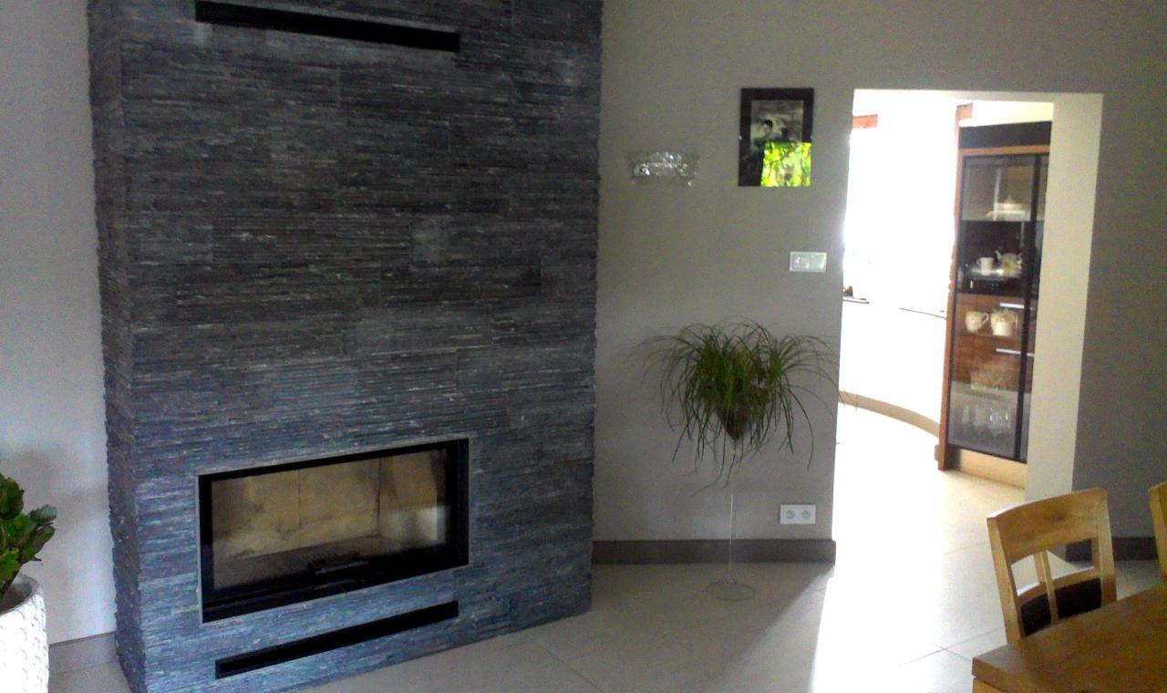 Nowoczesny kominek obudowany szarym kamieniem łupkowym, z widoczną ramą paleniska, na tle jasnego wnętrza z dekoracyjną rośliną doniczkową.