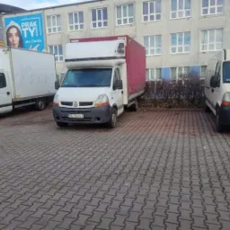 Trzy białe busy transportowe zaparkowane na brukowanym placu przed budynkiem z wieloma oknami, jeden z czerwonym dachem i reklamą na pobliskim budynku.