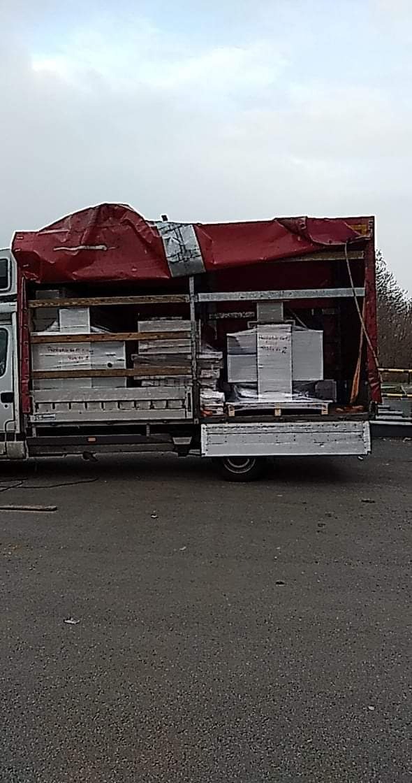 Ciężarówka z uszkodzoną czerwoną plandeką załadowana paletami z zapakowanymi kartonami, widok z tyłu na szarym asfalcie.