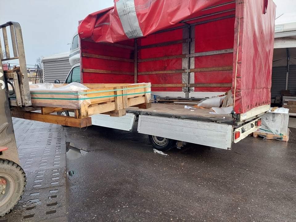 Wózek widłowy ładuje drewnianą skrzynię z towarem, owiniętą folią, na otwartą platformę dostawczego samochodu z czerwoną plandeką, widok z boku na mokry plac.