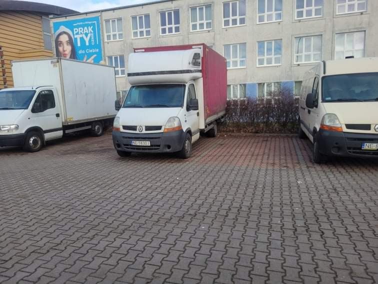 Trzy białe busy transportowe zaparkowane na brukowanym placu przed budynkiem z wieloma oknami, jeden z czerwonym dachem i reklamą na pobliskim budynku.