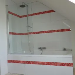 Wykończona łazienka na poddaszu: biała wanna z kabiną prysznicową, obłożona białymi płytkami z czerwonymi dekoracyjnymi pasami mozaiki. Ukośny sufit.