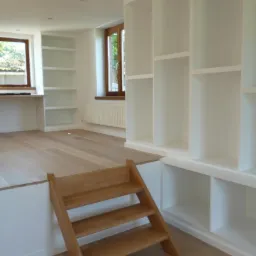 Jasne wnętrze z drewnianą podłogą i schodkami. Białe, wbudowane regały z otwartymi półkami. Dwa okna w drewnianych ramach w tle.