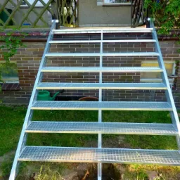 Zewnętrzne, metalowe schody kratowe prowadzą do wejścia domu z ceglaną ścianą. W tle zieleń ogrodu i okno piwniczne.