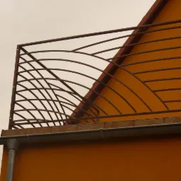 Metalowa balustrada balkonowa o dekoracyjnym, łukowatym wzorze, zamontowana na pomarańczowym budynku. Widok z dołu, podkreślający geometrię konstrukcji.