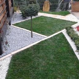 Landgreen projektowanie i zakładanie ogrodów - Geometryczny ogród w Lublinie: trawnik w kwadraty, białe i szare kamienie, drewniane ogrodzenie. Osoba podlewa rośliny w tle, donice z roślinami.