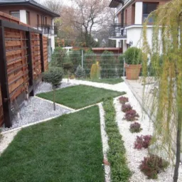 Ogród z trawnikiem, drewnianym płotem i ozdobnymi kamykami, drzewko płaczące. Geometryczne kształty trawnika i rabat. Dom w tle.