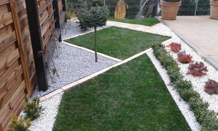 Geometryczny ogród w Lublinie: trawnik w kwadraty, białe i szare kamienie, drewniane ogrodzenie. Osoba podlewa rośliny w tle, donice z roślinami.