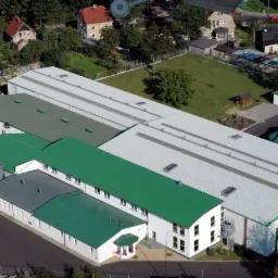 Widok z góry na kompleks budynków przemysłowych z zielonymi i srebrnymi dachami, otoczonych zielenią i zabudową mieszkalną w Gryfowie Śląskim.