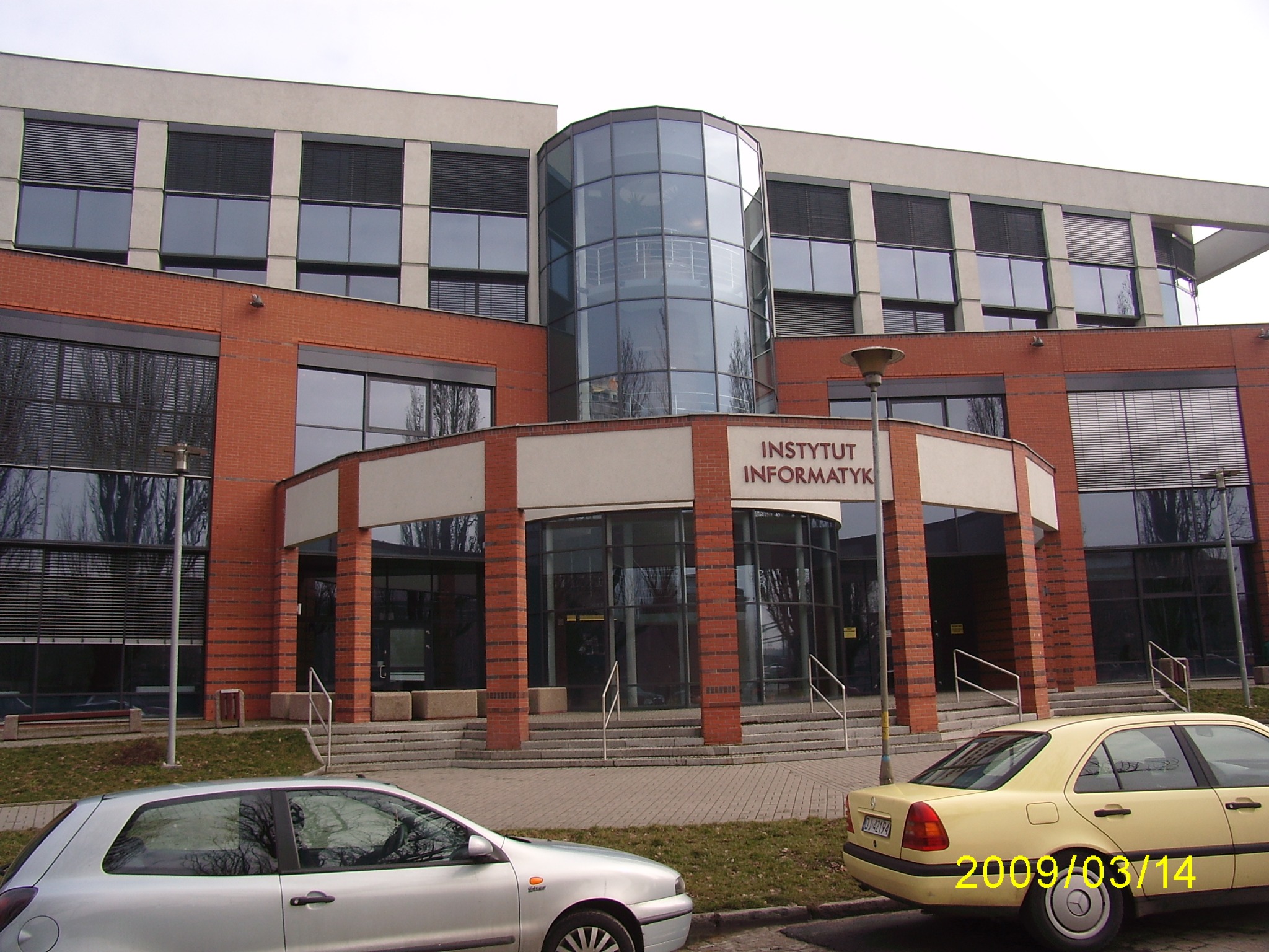 Budynek Instytutu Informatyki Uniwersytetu Wrocławskiego z charakterystycznym cylindrycznym, przeszklonym wejściem, wykonany z czerwonej cegły i jasnego tynku, z zaparkowanymi samochodami osobowymi...