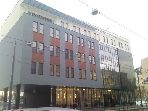 Budynek Starostwa Powiatowego we Wrocławiu z nowoczesną elewacją, łączącą szare panele, ceglane wstawki i duże okna, widziany z poziomu ulicy w pochmurny dzień.