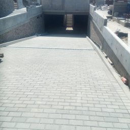 Zakład Remontowo Budowlany Jan Bednarz - Ułożona szara kostka brukowa na podjeździe do garażu podziemnego, z widocznym odpływem liniowym i fragmentem czerwonej kostki na dole kadru. W tle widoczne betonowe ściany i schody.