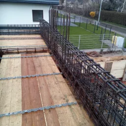 Zbrojenie pionowe z prętów stalowych w szalunku drewnianym na budowie domu, widok z góry na konstrukcję z elementami dystansowymi, w tle biały dom z szarą dachówką i zielony trawnik.