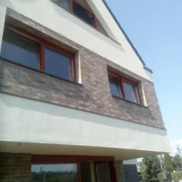 Nowoczesny dom z elewacją w odcieniach bieli i szarości, z fragmentem muru z cegły klinkierowej i oknami z ramami w kolorze mahoniu, widziany z dołu pod kątem.