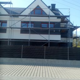 Zakład Remontowo Budowlany Jan Bednarz - Dom w trakcie ocieplania, widoczne rusztowania, częściowo ocieplona ściana w kolorze grafitowym, biała elewacja, okna z brązowymi ramami, siatka ogrodzeniowa, kostka brukowa na podjeździe...