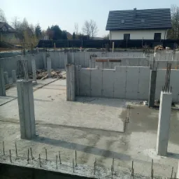 Surowe fundamenty domu z betonu, wystające pręty zbrojeniowe, widok na budynek mieszkalny w tle, wczesna faza budowy.