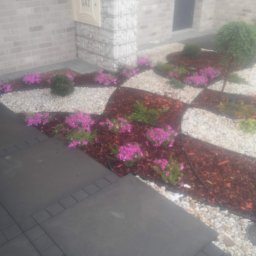 Zakład Remontowo Budowlany Jan Bednarz - Ogród przed domem z geometrycznym wzorem z białych kamieni, kory i różowych kwiatów, obok ścieżka z kostki brukowej i ściana budynku z licznikiem gazu.