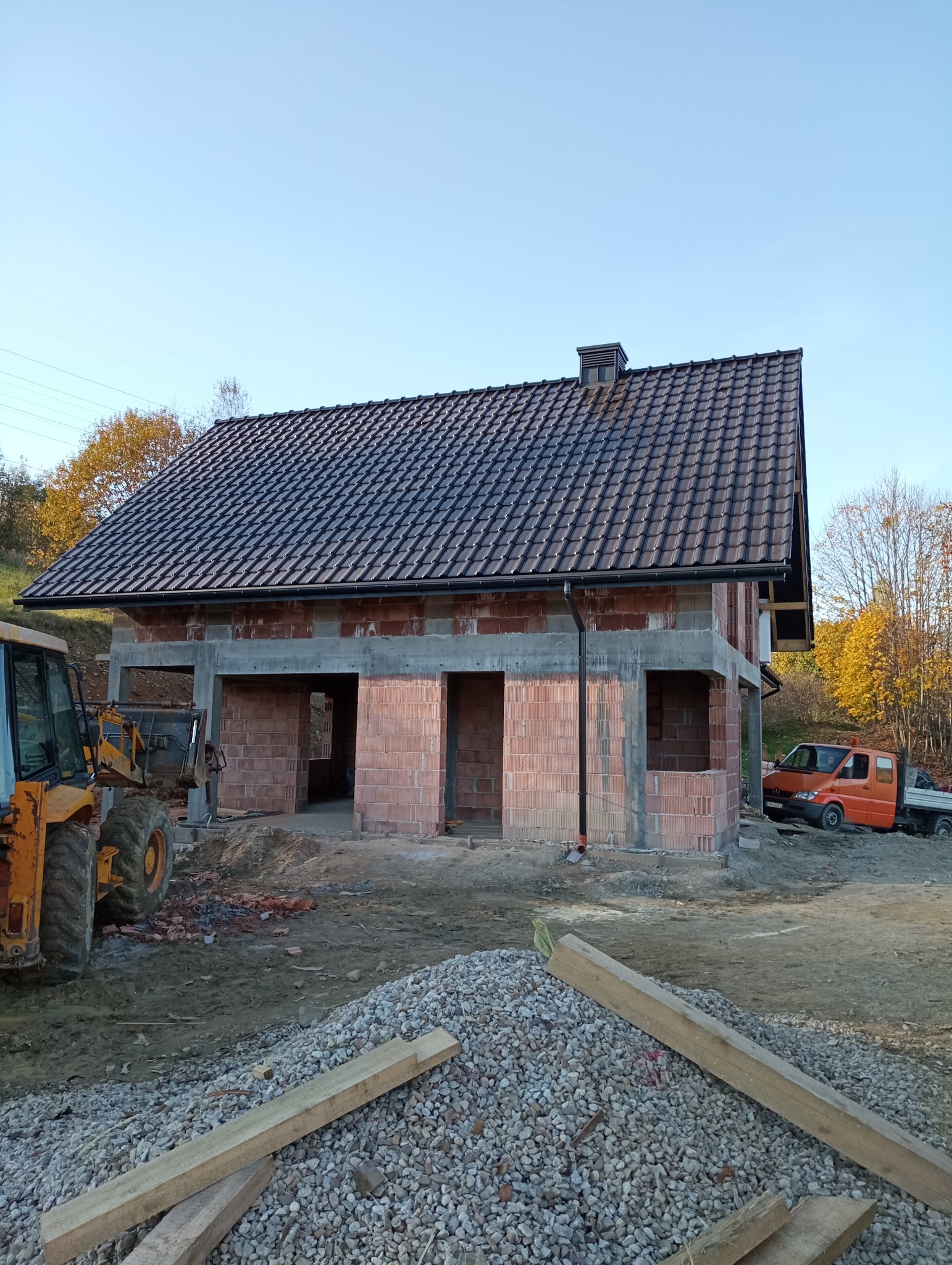 Surowy stan domu w trakcie budowy: ściany z czerwonej cegły, czarny dach z rynnami, widoczny kopiec żwiru i żółta koparka na tle jesiennego krajobrazu.