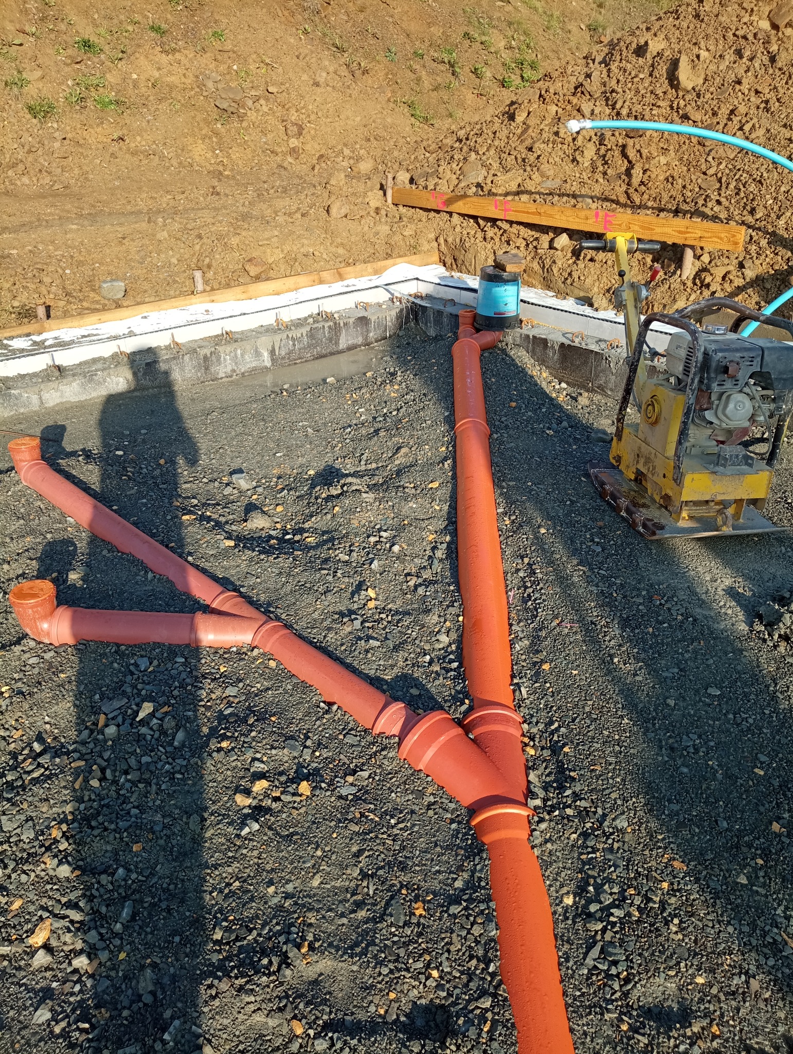Pomarańczowe rury kanalizacyjne w kształcie litery Y na żwirowym podłożu, z widocznym fundamentem budynku w tle i ubijarką wibracyjną.
