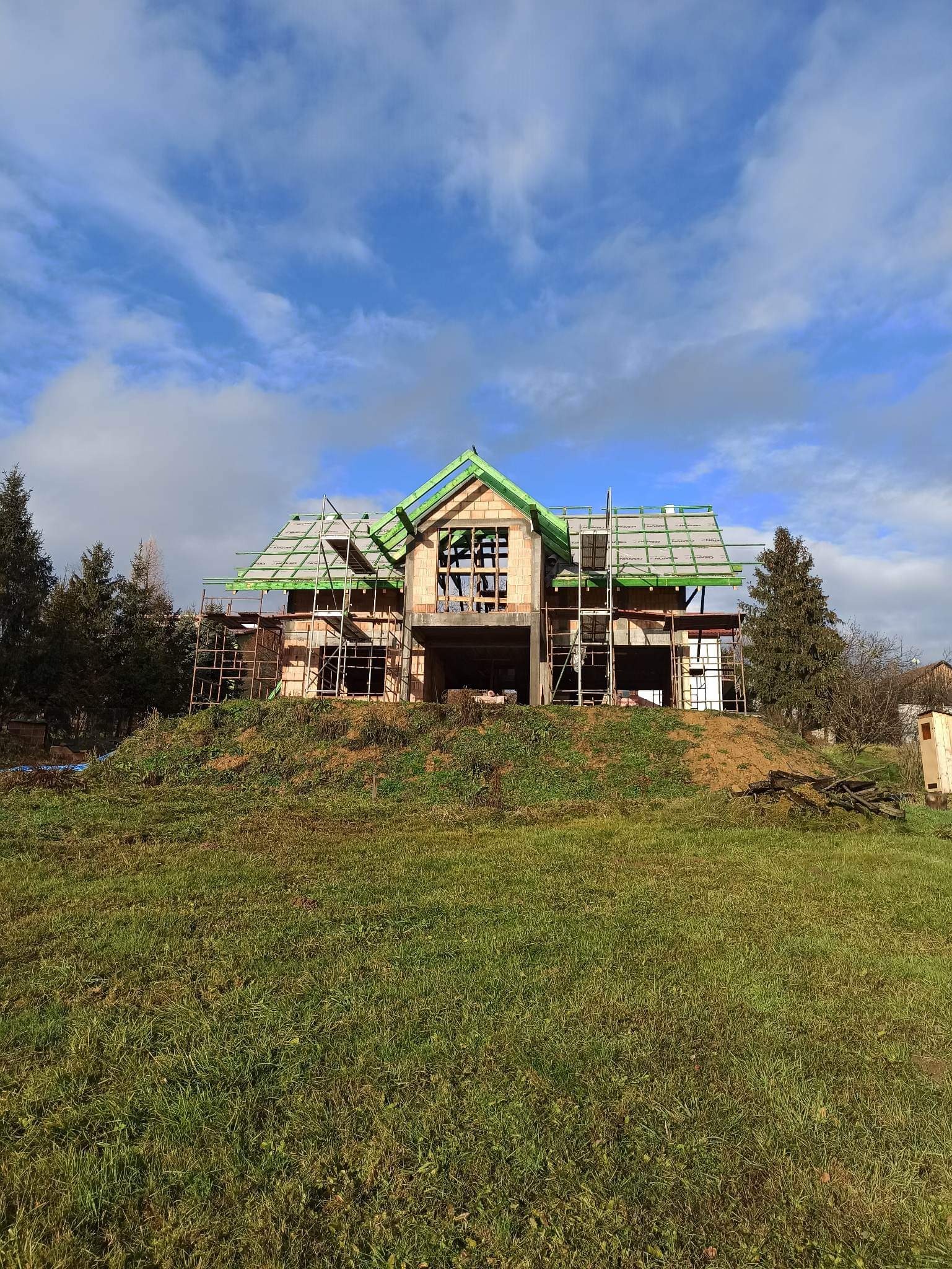 Surowy stan domu murowanego z cegły, z widocznym szkieletem dachu pokrytym zieloną folią i rusztowaniami, na tle zielonego wzgórza i błękitnego nieba z chmurami.