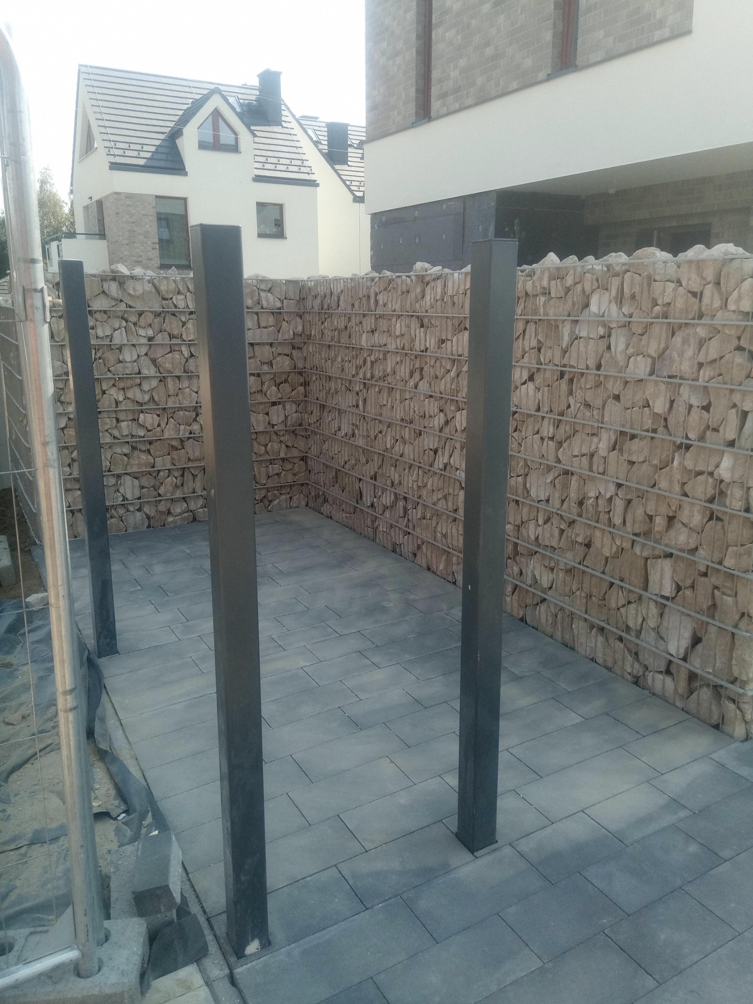 Gabionowa ściana oporowa z szarego kamienia, oddzielająca przestrzeń z szarą kostką brukową, z widocznymi metalowymi słupkami i częściowo widocznym domem w tle.