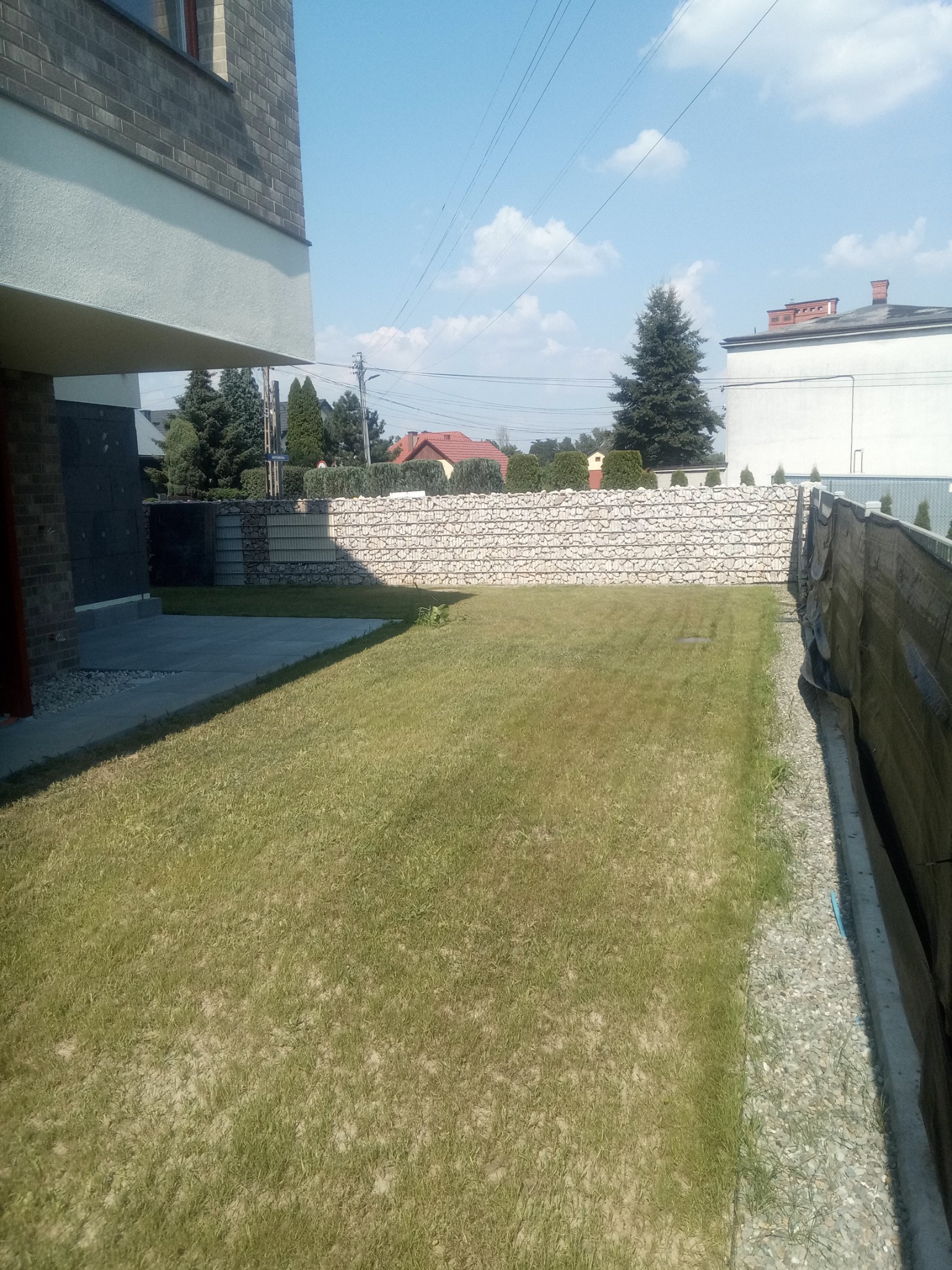 Widok na trawnik przed nowoczesnym domem z gabionowym ogrodzeniem, widoczne elementy elewacji budynku i sąsiednie domy w tle.