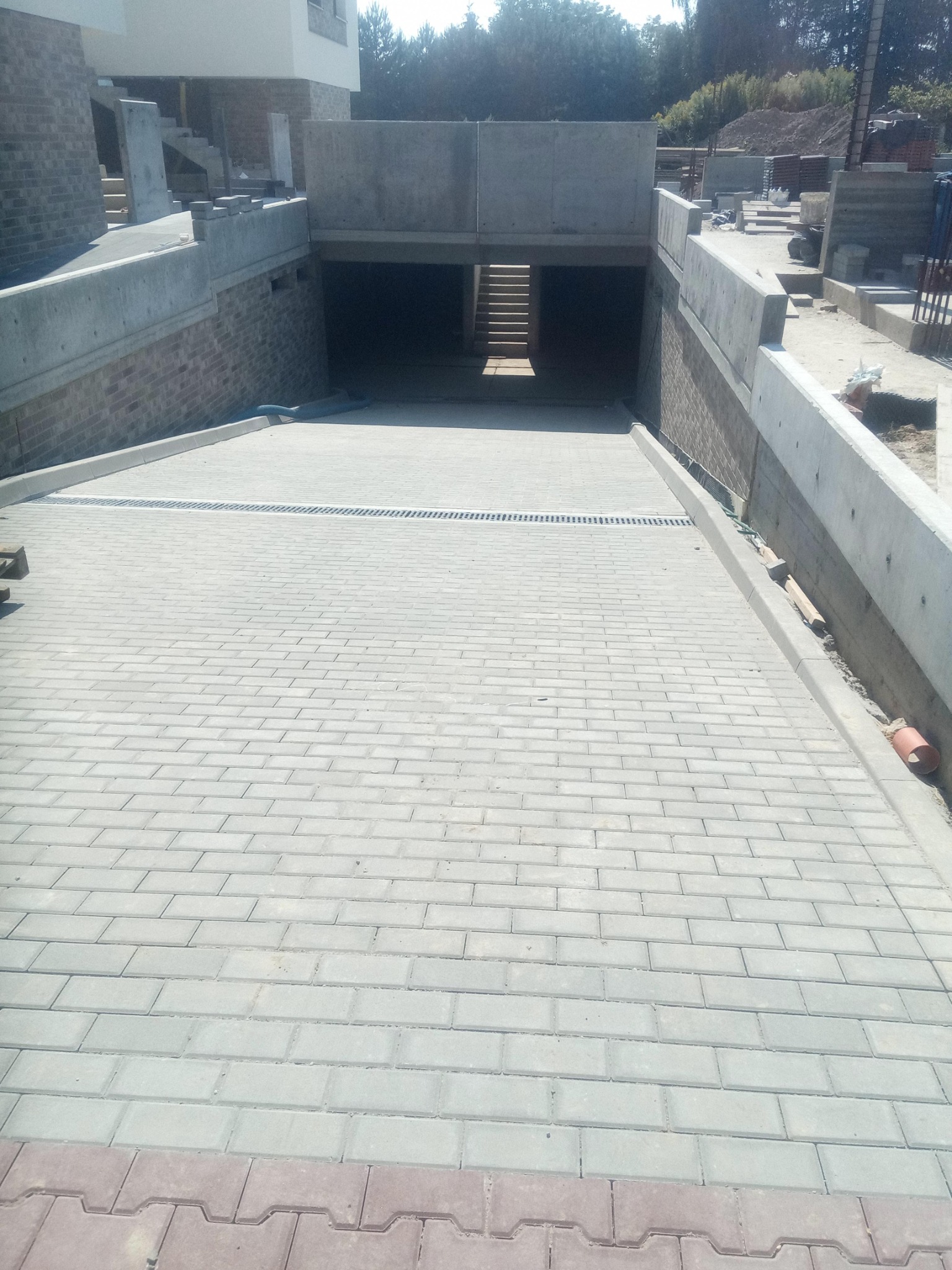 Ułożona szara kostka brukowa na podjeździe do garażu podziemnego, z widocznym odpływem liniowym i fragmentem czerwonej kostki na dole kadru. W tle widoczne betonowe ściany i schody.