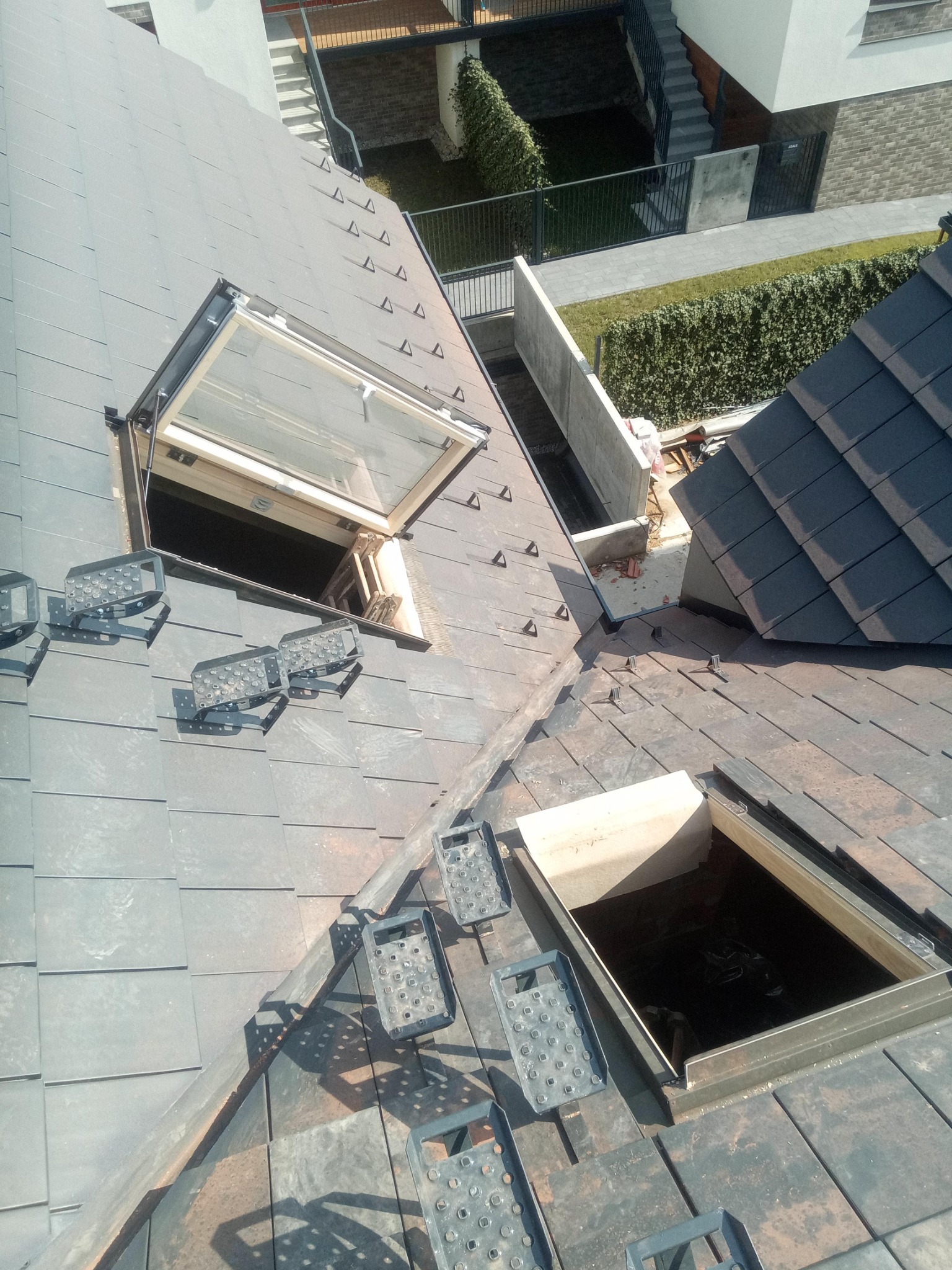 Montaż okna dachowego na dachu z ciemnej dachówki, widoczne stopnie dekarskie i otwarta rama okienna.