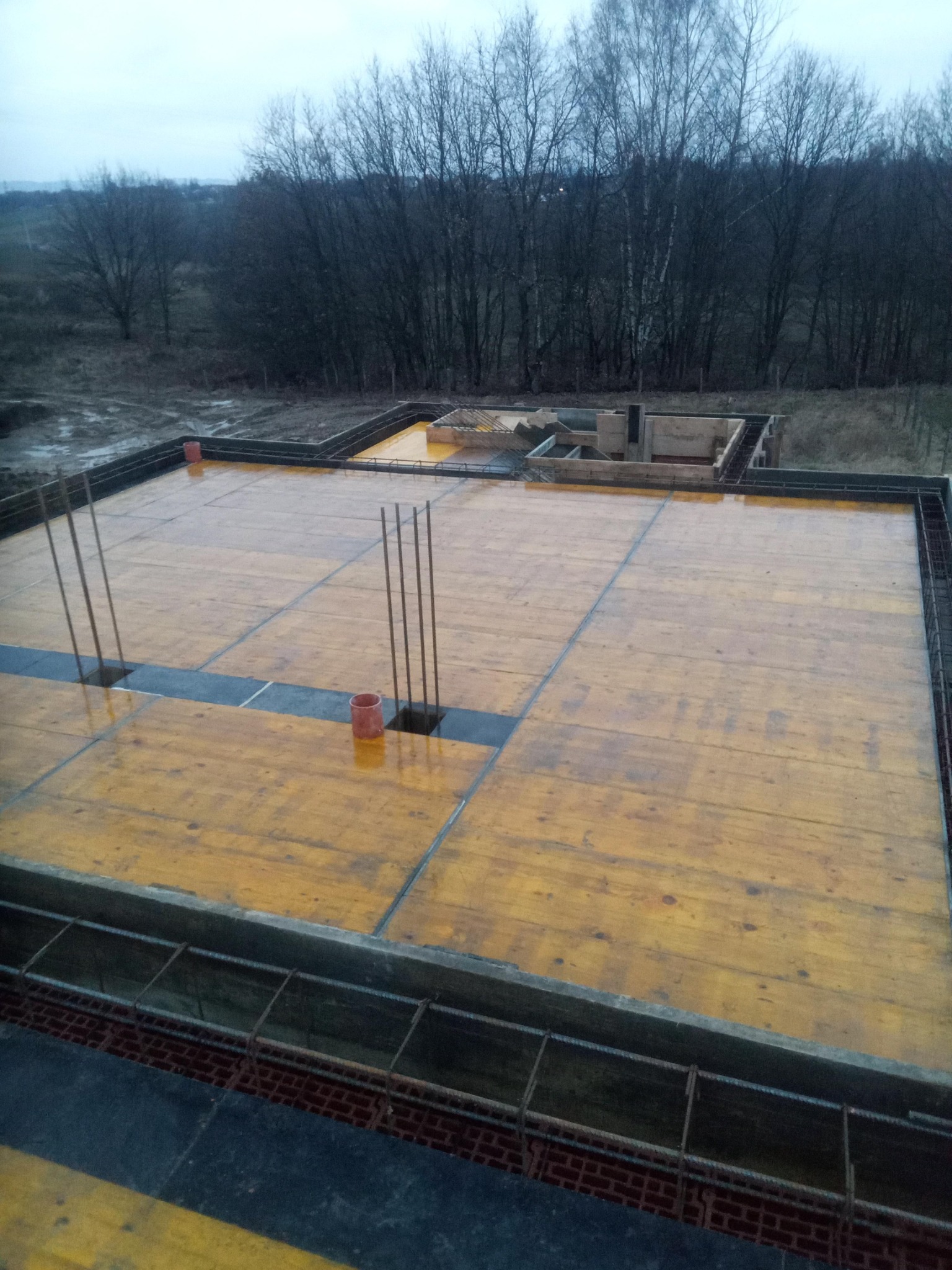 Deskowanie fundamentów domu jednorodzinnego z wystającymi prętami zbrojeniowymi, widok z góry na plac budowy w pochmurny dzień.