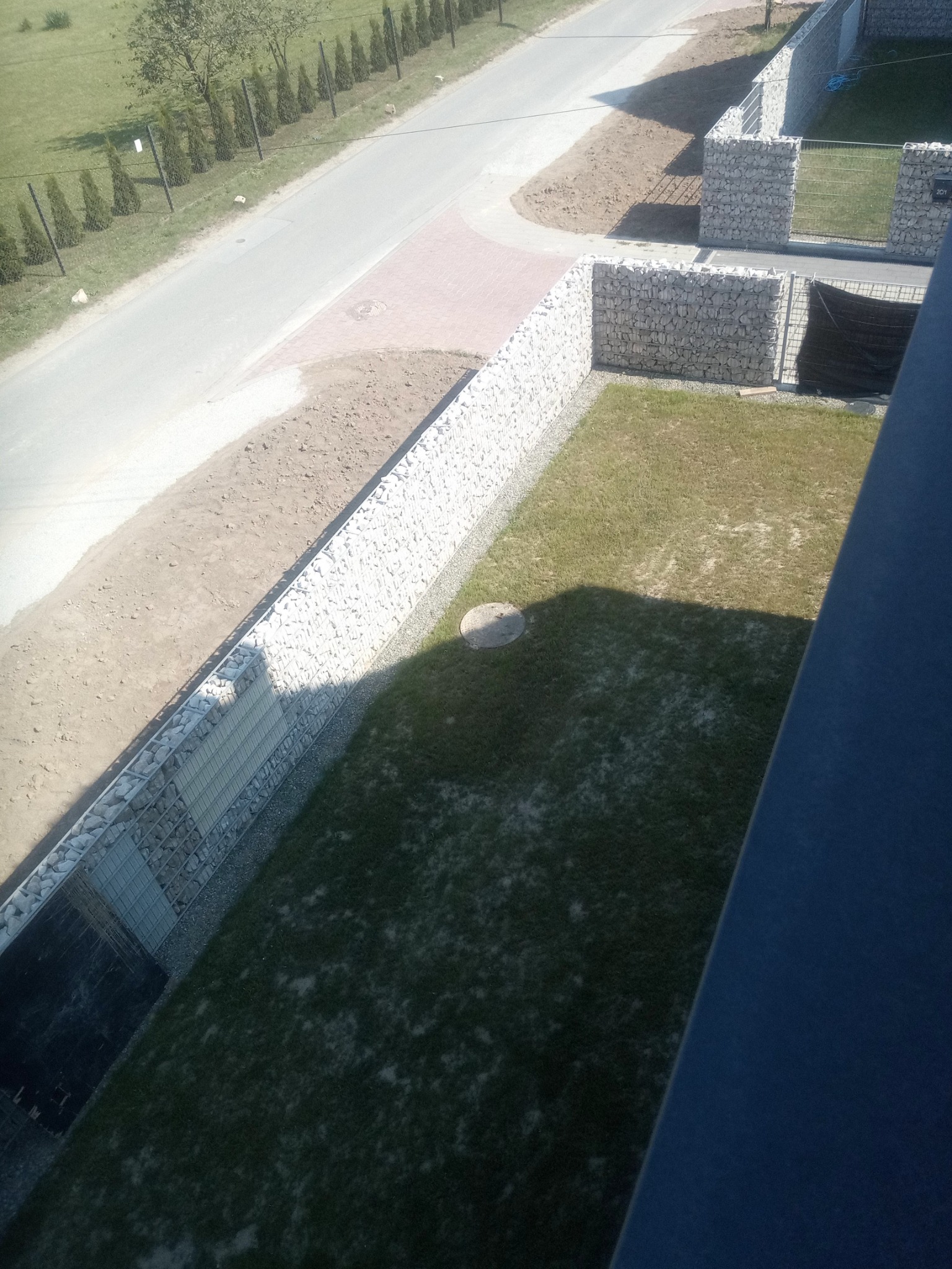 Gabionowe ogrodzenie z kamienia, widok z góry na posesję z trawnikiem, drogą i rzędem drzew w tle.