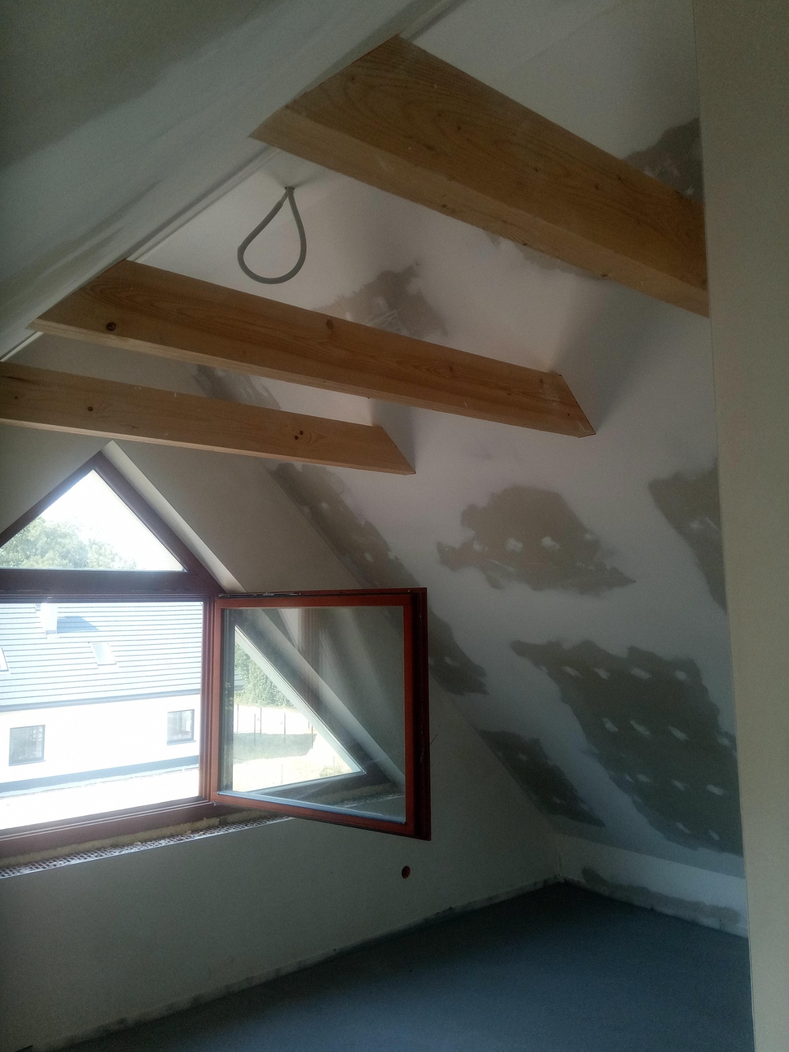 Poddasze w trakcie wykańczania: drewniane belki stropowe, okno dachowe z widokiem na dom, ściany z widocznymi łatami gipsowymi przed malowaniem.